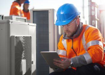 An air conditioning engineer is finishing the installation of several units on a rooftop. Two colleagues can be seen also installing units in the background. They are wearing hi vis jackets, hard hats and safety goggles.