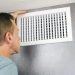 Mature man examining an outflow air vent grid and duct to see if it needs cleaning. One guy looking into a home air duct to see how clean and healthy it is.