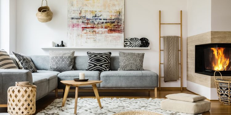 Wooden table next to grey corner settee in warm living room interior with painting and fireplace. Real photo