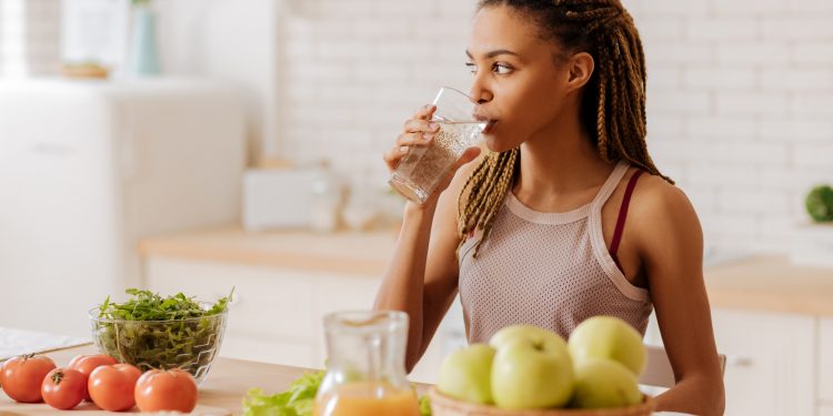 Water before breakfast. Slim and fit woman with many little braids drinking water before having breakfast