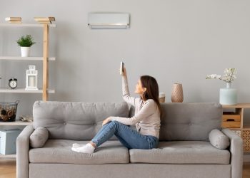 Young smiling woman in casual wear sitting on couch in living room resting holding remote controller switch on air conditioner make regulate comfort temperature at home enjoy fresh or warm air concept.