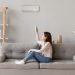Young smiling woman in casual wear sitting on couch in living room resting holding remote controller switch on air conditioner make regulate comfort temperature at home enjoy fresh or warm air concept.