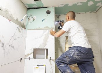 Plumber using electric screwdriver. Bathroom renovation concept. Marble ceramic tiles with spacers and grey cement walls in bathroom. Industrial builder working in toilet