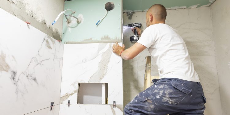 Plumber using electric screwdriver. Bathroom renovation concept. Marble ceramic tiles with spacers and grey cement walls in bathroom. Industrial builder working in toilet