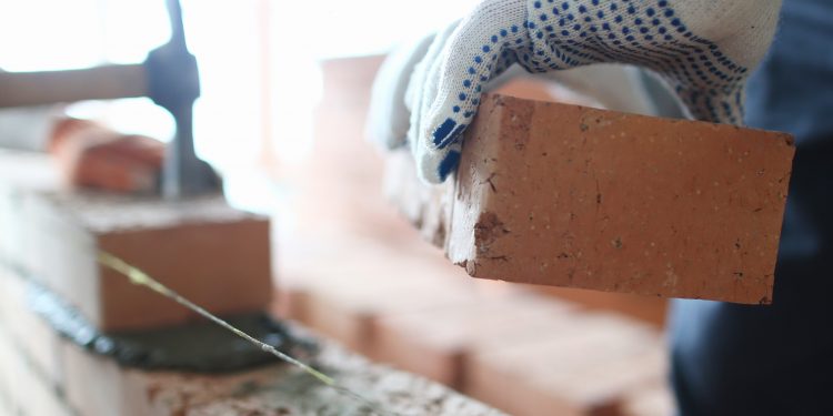 Male builder hand in gloves holding clay brick on background of brickwork wall. Repair and redevelopment apartments concept.