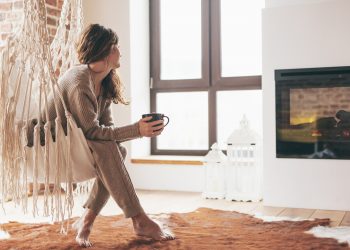 Beautiful young woman, which is wearing cashmere nightwear clothes, drinking tea or coffee while relaxing on rocking chair in scandinavian cabin near electric heater fireplace one lazy weekend morning