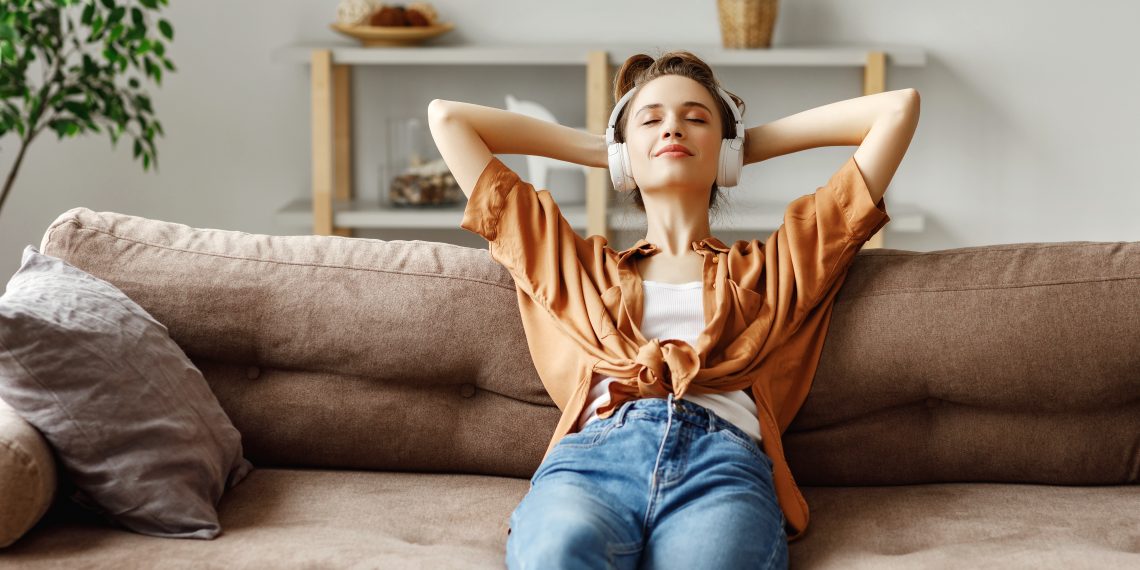 Happy millennial female with eyes closed in wireless headphones and casual clothes sitting alone on soft couch with hands behind head and listening with pleasure to favorite music in light spacious living room