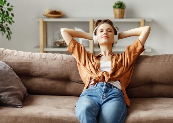 Happy millennial female with eyes closed in wireless headphones and casual clothes sitting alone on soft couch with hands behind head and listening with pleasure to favorite music in light spacious living room