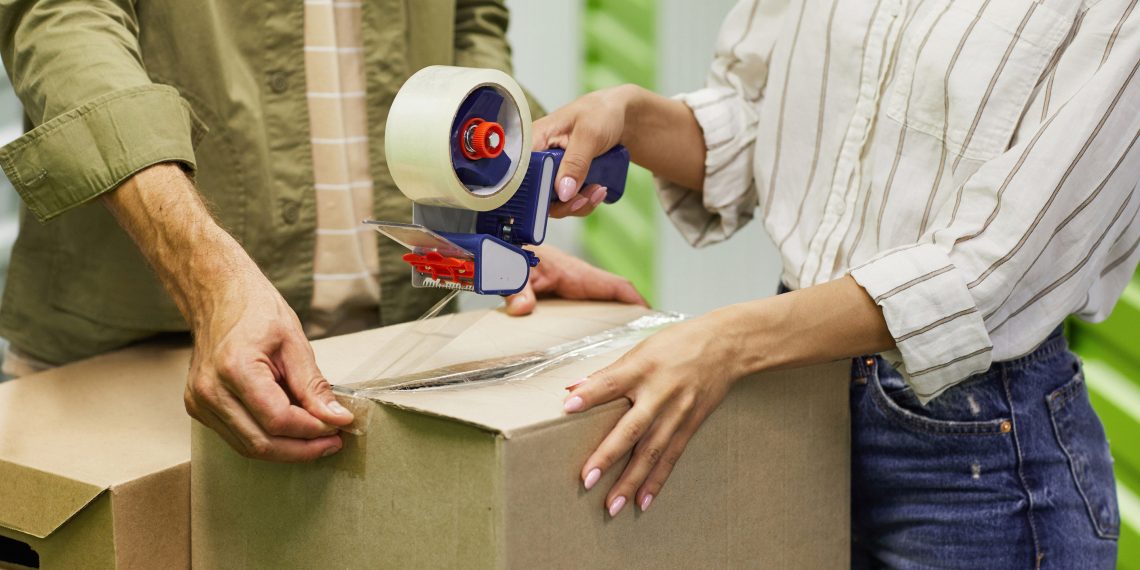 Close up of unrecognizable couple packing boxes with tape gun while standing in self storage unit, copy space