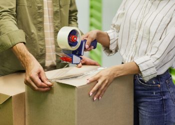 Close up of unrecognizable couple packing boxes with tape gun while standing in self storage unit, copy space