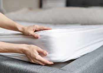 Cropped view of woman hand checking quality and putting new orthopedic mattress on sleeping bed in bedroom. Female demonstrating soft hypoallergenic foam. Maid changes bedding linen in the hotel room