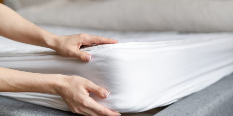 Cropped view of woman hand checking quality and putting new orthopedic mattress on sleeping bed in bedroom. Female demonstrating soft hypoallergenic foam. Maid changes bedding linen in the hotel room