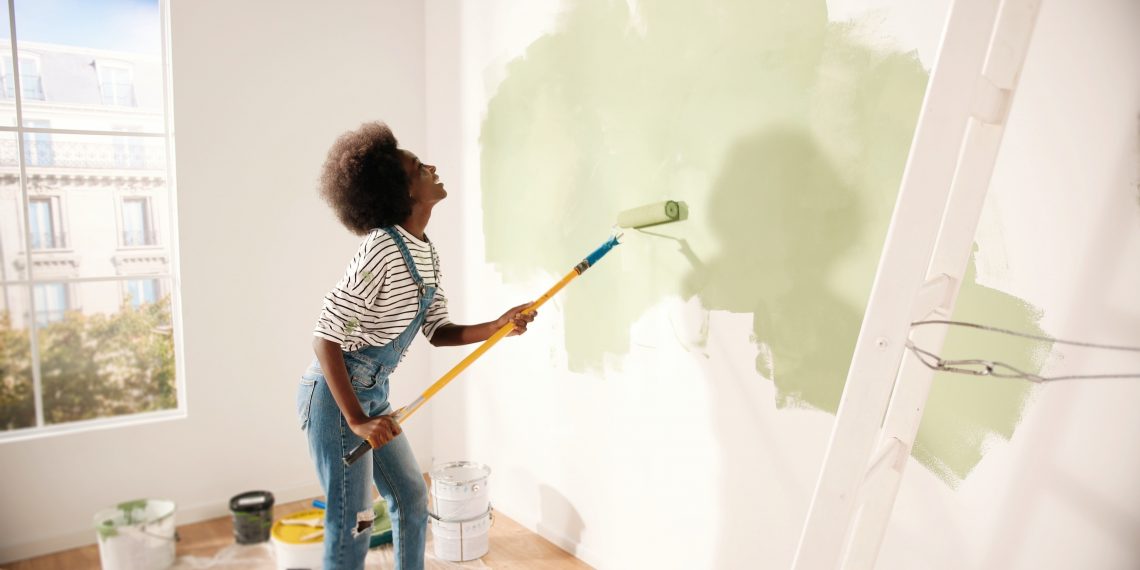 Young African American woman dancing and painting wall with roller brush while renovating apartment. Rear of female having fun redecorating home, renovating and improving Repair and decorating concept