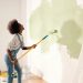 Young African American woman dancing and painting wall with roller brush while renovating apartment. Rear of female having fun redecorating home, renovating and improving Repair and decorating concept