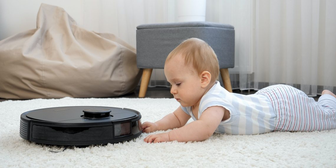 Cut elittle baby lying on carpet at house and looking at robot vacuum cleaner doing cleanup on carpet.