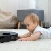 Cut elittle baby lying on carpet at house and looking at robot vacuum cleaner doing cleanup on carpet.