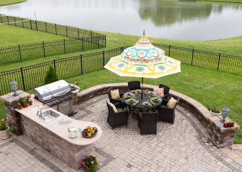 Outdoor living space on a brick patio overlooking a tranquil lake and fenced green lawn with a table under a sunshade or umbrella laid ready for dinner, high angle view