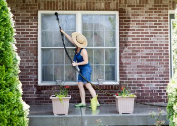 Housewife standing on a patio washing the windows of her house with a hose attachment as she spring-cleans the exterior at the start of the new spring season