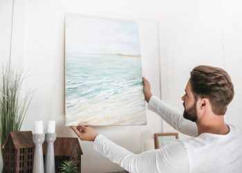 Young bearded man hanging picture on wall at home