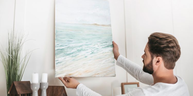 Young bearded man hanging picture on wall at home