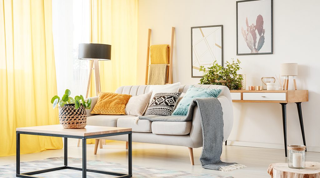 Plant on table and lamp in warm living room with yellow curtains, posters and pillows on sofa
